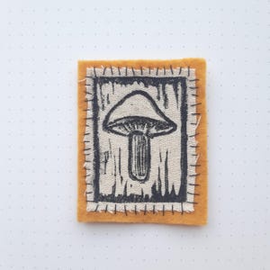May include: A brown felt patch with a black stitched border. The patch features a black and white illustration of a mushroom on a white background.