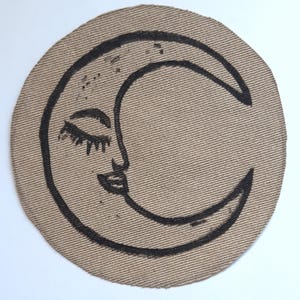 May include: A circular tan fabric patch featuring a black embroidered crescent moon design. The moon has a stylised face with closed eyes, long eyelashes, and a small mouth, rendered in bold black thread against the textured tan background.