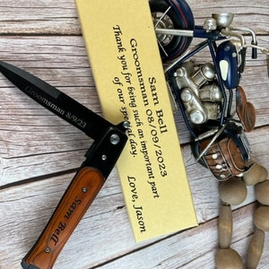Peut inclure: Un couteau pliant noir avec un manche en bois, gravé de "Sam Bell" et "Groomsman 8/9/23." Une petite maquette de moto décorative et une carte avec du texte sont également présentes. La carte indique "Groomsman 08/09/2023" et "Merci d'avoir joué un rôle si important dans notre journée spéciale."