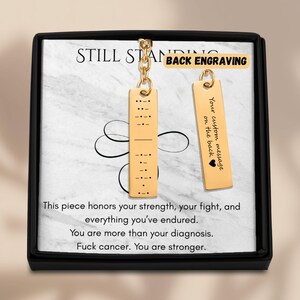 Peut inclure: Deux porte-clés pendentifs dorés avec du code Morse et le texte "Your custom message on the back". Les porte-clés sont présentés dans une boîte noire avec le texte "STILL STANDING" et un message sur la force et le cancer.