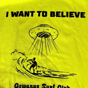 May include: Bright yellow t-shirt with the text "I WANT TO BELIEVE" above a graphic of a UFO beaming down on a surfer. Below the graphic is "Gowanus Surf Club Brooklyn NYC" in black text.