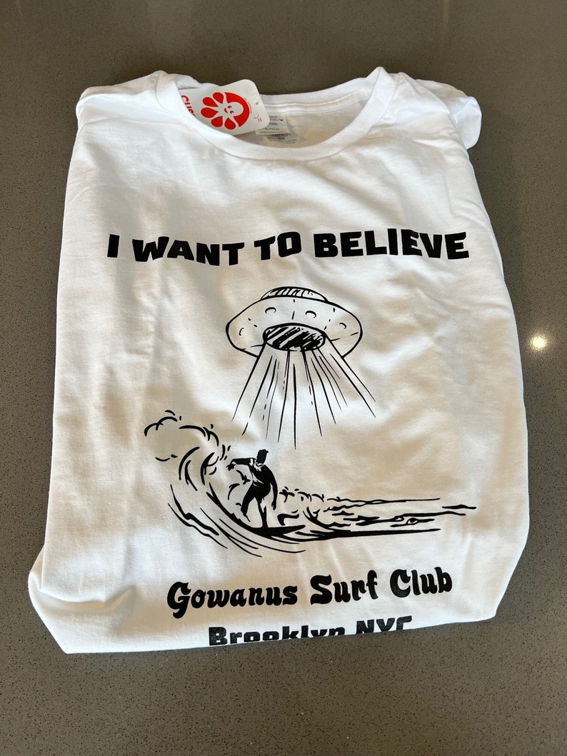 May include: White t-shirt with the text "I WANT TO BELIEVE" above a black illustration of a UFO and a surfer riding a wave. The shirt also features "Gowanus Surf Club" and "Brooklyn NYC" in black text.