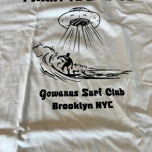 May include: White t-shirt with a black graphic of a UFO beaming down on a surfer riding a wave. Text above reads "I WANT TO BELIEVE", with "Gowanus Surf Club Brooklyn NYC" below.
