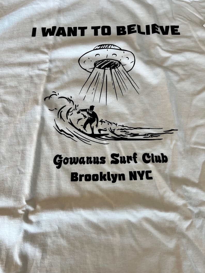 May include: White t-shirt with a black graphic of a UFO beaming down on a surfer riding a wave. Text above reads "I WANT TO BELIEVE", with "Gowanus Surf Club Brooklyn NYC" below.