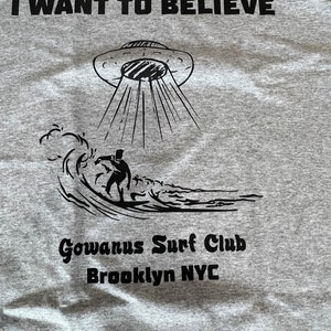 May include: Gray t-shirt with the text "I WANT TO BELIEVE" above a UFO graphic beaming down on a surfer riding a wave. Below the graphic is "Gowanus Surf Club Brooklyn NYC".