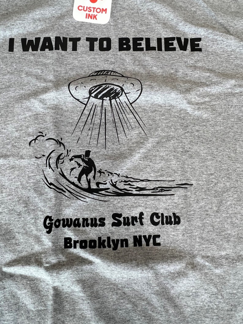 May include: Gray t-shirt with the text "I WANT TO BELIEVE" above a UFO graphic beaming down on a surfer riding a wave. Below the graphic is "Gowanus Surf Club Brooklyn NYC".