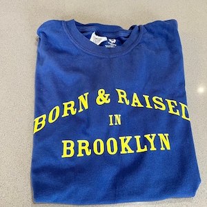 以下が含まれることがあります： 「BORN & RAISED IN BROOKLYN」という黄色の文字が書かれた青いTシャツです。