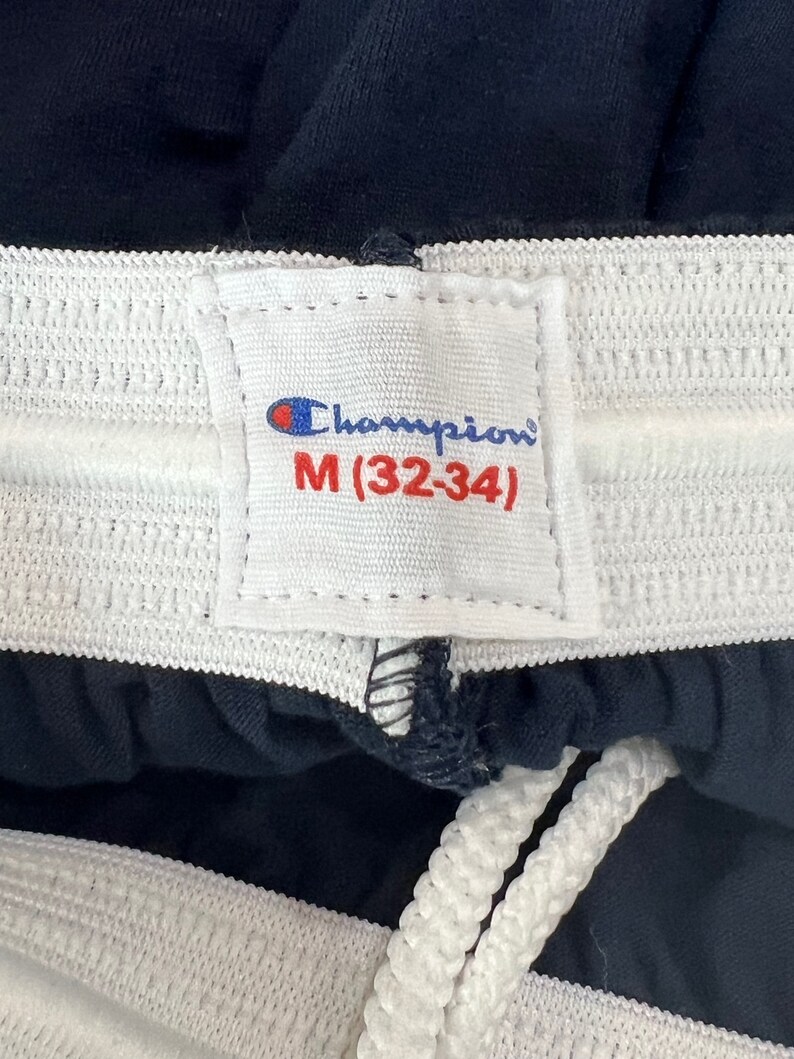 May include: A white tag with black stitching and the text "Champion" and "M (32-34)" on a navy blue fabric background.