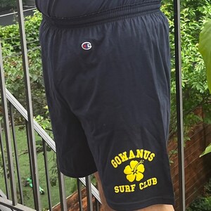 May include: Black athletic shorts with a yellow logo that says "Gowanus Surf Club" and a yellow hibiscus flower. The shorts have a white Champion logo on the back.