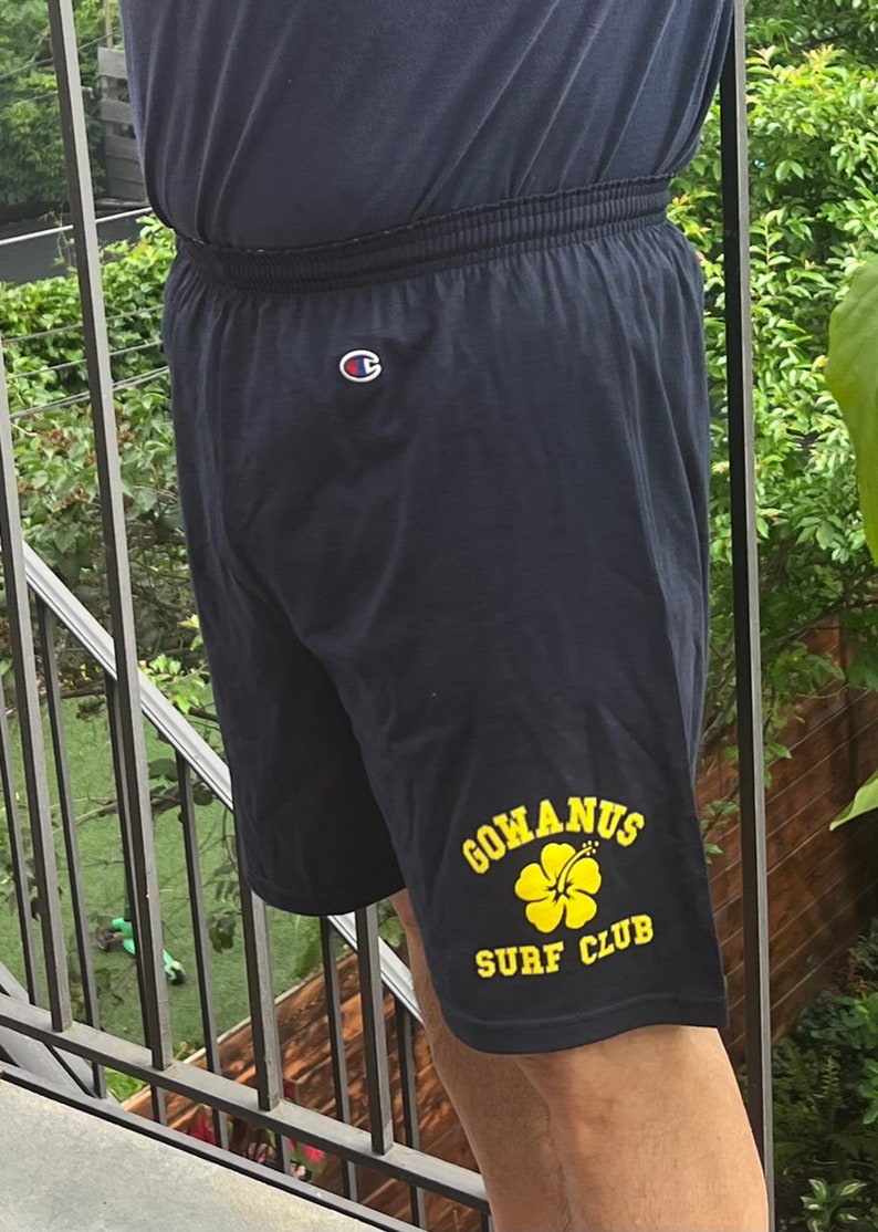 May include: Black athletic shorts with a yellow logo that says "Gowanus Surf Club" and a yellow hibiscus flower. The shorts have a white Champion logo on the back.