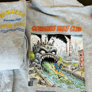 以下が含まれることがあります： グレーのフード付きスウェットシャツ。モンスターと街のシルエットが描かれたグラフィックプリントが施されています。スウェットシャツの前にはオレンジ色の「GOWANUS SURF CLUB」の文字がプリントされています。グラフィックの下にはオレンジ色の「BROOKLYN, NEW YORK」の文字がプリントされています。