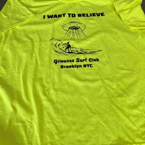 May include: Neon yellow t-shirt with black text and graphics. The text reads "I WANT TO BELIEVE" above an illustration of a UFO and a surfer. Below the graphic is "Gowanus Surf Club Brooklyn NYC".