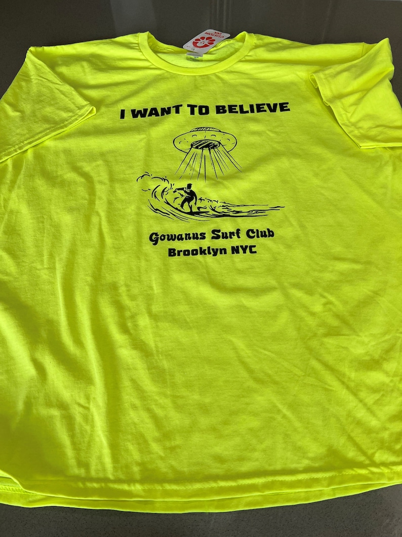 May include: Neon yellow t-shirt with black text and graphics. The text reads "I WANT TO BELIEVE" above an illustration of a UFO and a surfer. Below the graphic is "Gowanus Surf Club Brooklyn NYC".