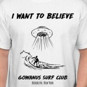 May include: White t-shirt with a black graphic featuring a UFO, a surfer, and the text "I WANT TO BELIEVE" above and "GOWANUS SURF CLUB BROOKLYN, NEW YORK" below. The design is a line drawing.