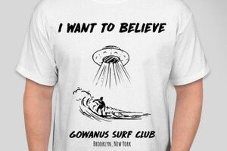 May include: White t-shirt with a black graphic featuring a UFO, a surfer, and the text "I WANT TO BELIEVE" above and "GOWANUS SURF CLUB BROOKLYN, NEW YORK" below. The design is a line drawing.