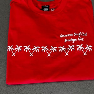 May include: Red t-shirt with a white palm tree design and the text "Gowanus Surf Club Brooklyn NYC."