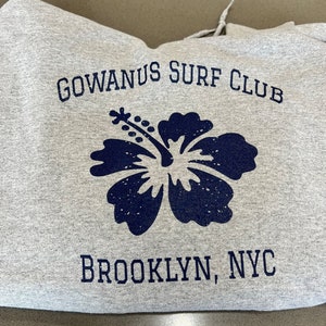 以下が含まれることがあります： ハイビスカスの花と「GOWANUS SURF CLUB」と「BROOKLYN, NYC」の文字が青いプリントされたグレーのフード付きスウェットシャツ。