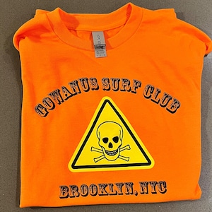 以下が含まれることがあります： 黄色い三角形の中に、黒い骸骨と交差した骨のグラフィックが描かれたオレンジ色のTシャツ。三角形の上に「GOWANUS SURF CLUB」という文字が、下に「BROOKLYN, NYC」という文字が書かれている。