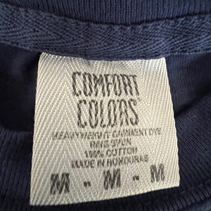 May include: A white tag with black text that reads "COMFORT COLORS" and "HEAVYWEIGHT GARMENT DYE", "RAVE SPUN", "100% COTTON", "MADE IN HONDURAS", and "M - M - M".
