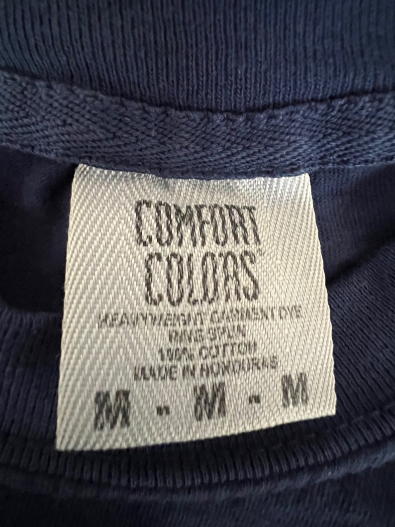 May include: A white tag with black text that reads "COMFORT COLORS" and "HEAVYWEIGHT GARMENT DYE", "RAVE SPUN", "100% COTTON", "MADE IN HONDURAS", and "M - M - M".