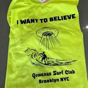 May include: Neon yellow t-shirt with the phrase "I WANT TO BELIEVE" above a graphic of a UFO beaming down on a surfer. The shirt also features "Gowanus Surf Club Brooklyn NYC" in black text.