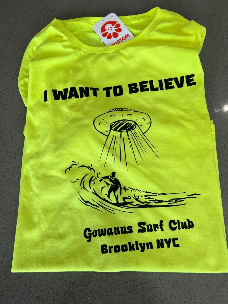 May include: Neon yellow t-shirt with the phrase "I WANT TO BELIEVE" above a graphic of a UFO beaming down on a surfer. The shirt also features "Gowanus Surf Club Brooklyn NYC" in black text.