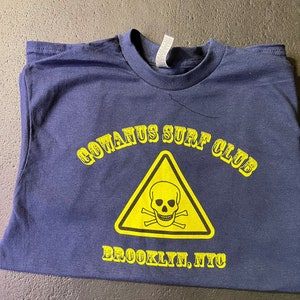 以下が含まれることがあります： 黄色のグラフィックが施されたネイビーブルーのTシャツ。三角形の中にドクロとクロスボーンが描かれており、その下に「GOWANUS SURF CLUB」と「BROOKLYN, NYC」の文字が書かれている。