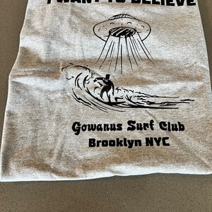 May include: A gray t-shirt with the words "I WANT TO BELIEVE" above a black illustration of a UFO beaming down on a surfer riding a wave. Below is "Gowanus Surf Club Brooklyn NYC".