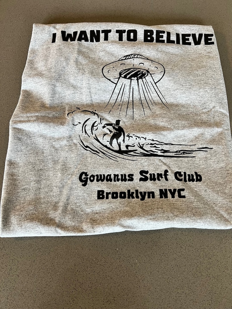 May include: A gray t-shirt with the words "I WANT TO BELIEVE" above a black illustration of a UFO beaming down on a surfer riding a wave. Below is "Gowanus Surf Club Brooklyn NYC".