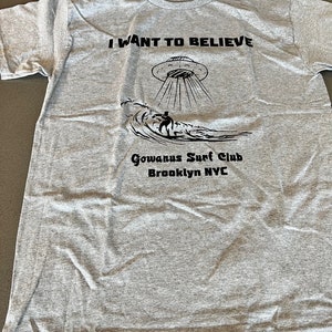 May include: Gray t-shirt with the text "I WANT TO BELIEVE" above a graphic of a UFO and a surfer on a wave. Below the graphic is "Gowanus Surf Club Brooklyn NYC".