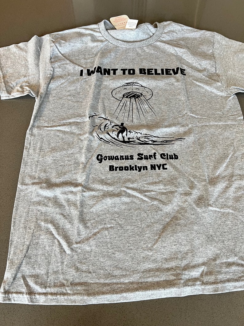 May include: Gray t-shirt with the text "I WANT TO BELIEVE" above a graphic of a UFO and a surfer on a wave. Below the graphic is "Gowanus Surf Club Brooklyn NYC".