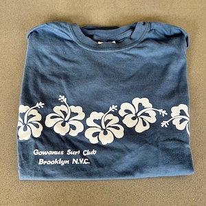 May include: A blue t-shirt with a white hibiscus flower design and the text "Gowanus Surf Club Brooklyn N.Y.C."