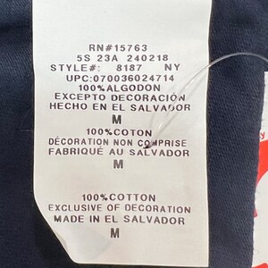 May include: A white tag with black text detailing the size, style, UPC, material, and origin of the garment. The tag reads "RN#15763 5S 23A 240218 STYLE#: 8187 NY UPC:070036024714 100% ALGODON EXCEPTO DECORACI&Oacute;N HECHO EN EL SALVADOR M 100% COTON D&Eacute;CORATION NON COMPRISE FABRIQU&Eacute; AU SALVADOR M 100% COTTON EXCLUSIVE OF DECORATION MADE IN EL SALVADOR M".