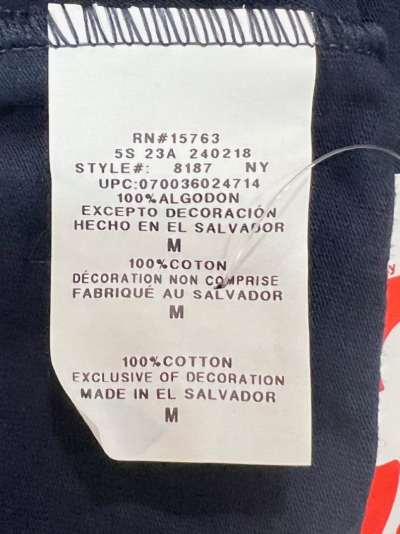 May include: A white tag with black text detailing the size, style, UPC, material, and origin of the garment. The tag reads "RN#15763 5S 23A 240218 STYLE#: 8187 NY UPC:070036024714 100% ALGODON EXCEPTO DECORACI&Oacute;N HECHO EN EL SALVADOR M 100% COTON D&Eacute;CORATION NON COMPRISE FABRIQU&Eacute; AU SALVADOR M 100% COTTON EXCLUSIVE OF DECORATION MADE IN EL SALVADOR M".
