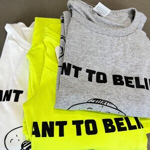 May include: Three folded t-shirts in white, neon yellow, and gray. Each shirt has the phrase "I WANT TO BELIEVE" printed in black, along with a cartoon UFO graphic. The shirts are stacked on a gray surface.