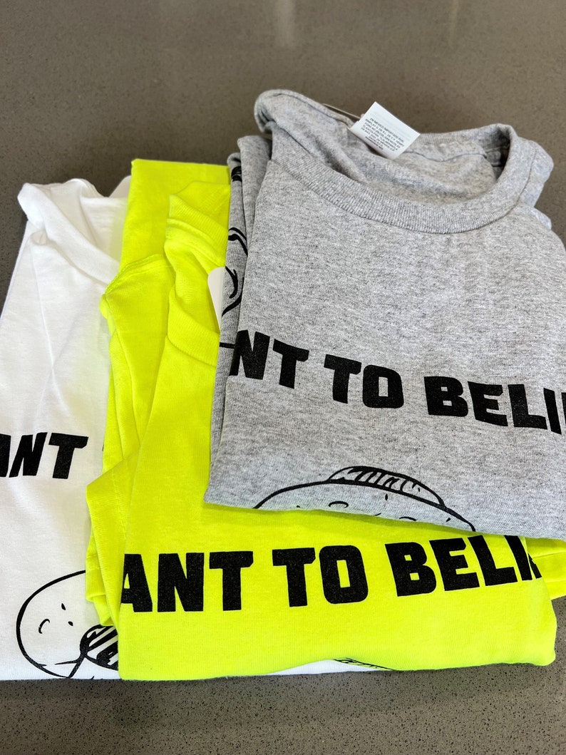 May include: Three folded t-shirts in white, neon yellow, and gray. Each shirt has the phrase "I WANT TO BELIEVE" printed in black, along with a cartoon UFO graphic. The shirts are stacked on a gray surface.