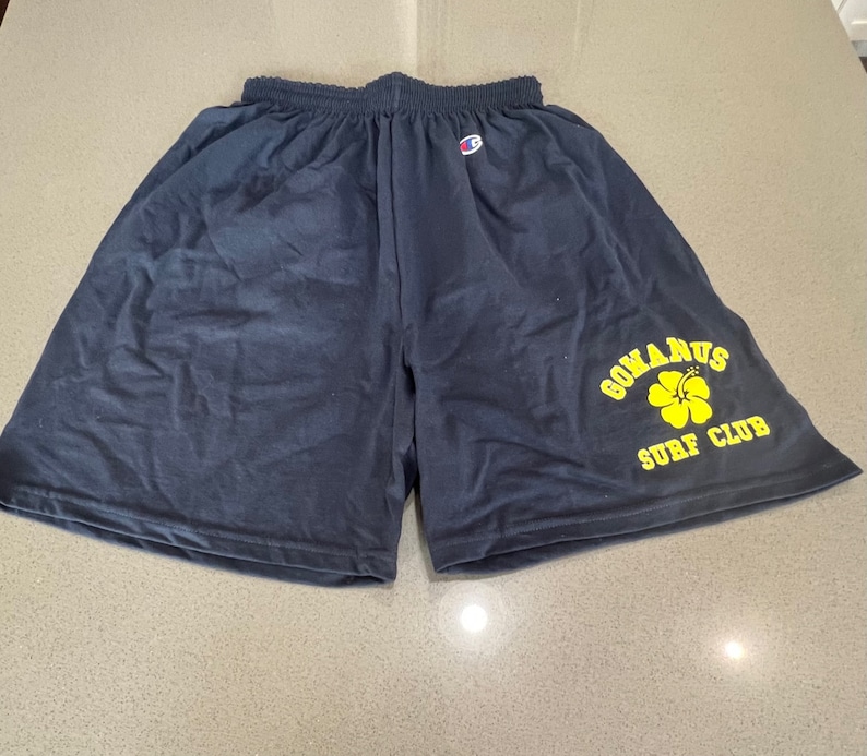 May include: Black athletic shorts with a yellow logo that says "GOMANUS SURF CLUB" and a yellow hibiscus flower.