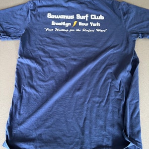 May include: A navy blue t-shirt with the text "Gowanus Surf Club Brooklyn New York" and "Just Waiting for the Perfect Wave" printed on the back.