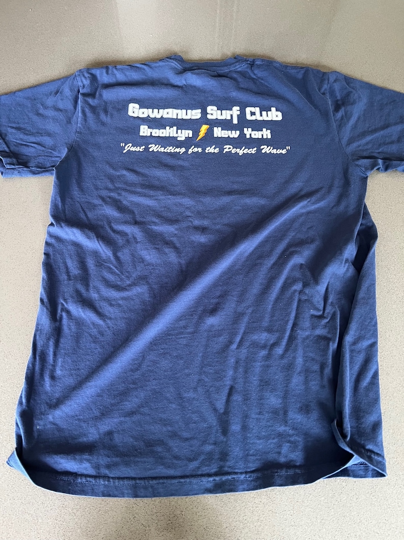 May include: A navy blue t-shirt with the text "Gowanus Surf Club Brooklyn New York" and "Just Waiting for the Perfect Wave" printed on the back.