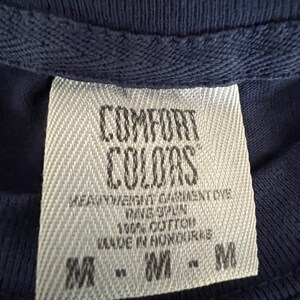 May include: A white tag with black text that reads "COMFORT COLORS" and "HEAVYWEIGHT GARMENT DYE", "RING SPUN", "100% COTTON", "MADE IN HONDURAS", and "M - M - M".