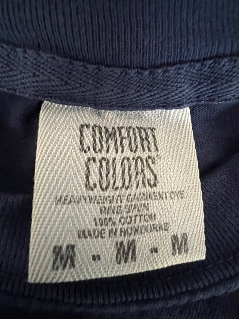 May include: A white tag with black text that reads "COMFORT COLORS" and "HEAVYWEIGHT GARMENT DYE", "RING SPUN", "100% COTTON", "MADE IN HONDURAS", and "M - M - M".