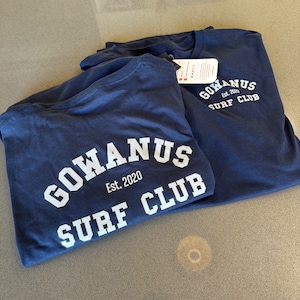 以下が含まれることがあります： 白い文字で「GOWANUS Est. 2020 SURF CLUB」と書かれた、紺色のTシャツが2枚です。