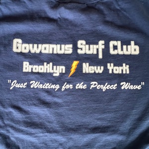 May include: A navy blue t-shirt with white text that reads "Gowanus Surf Club Brooklyn New York" and "Just Waiting for the Perfect Wave".