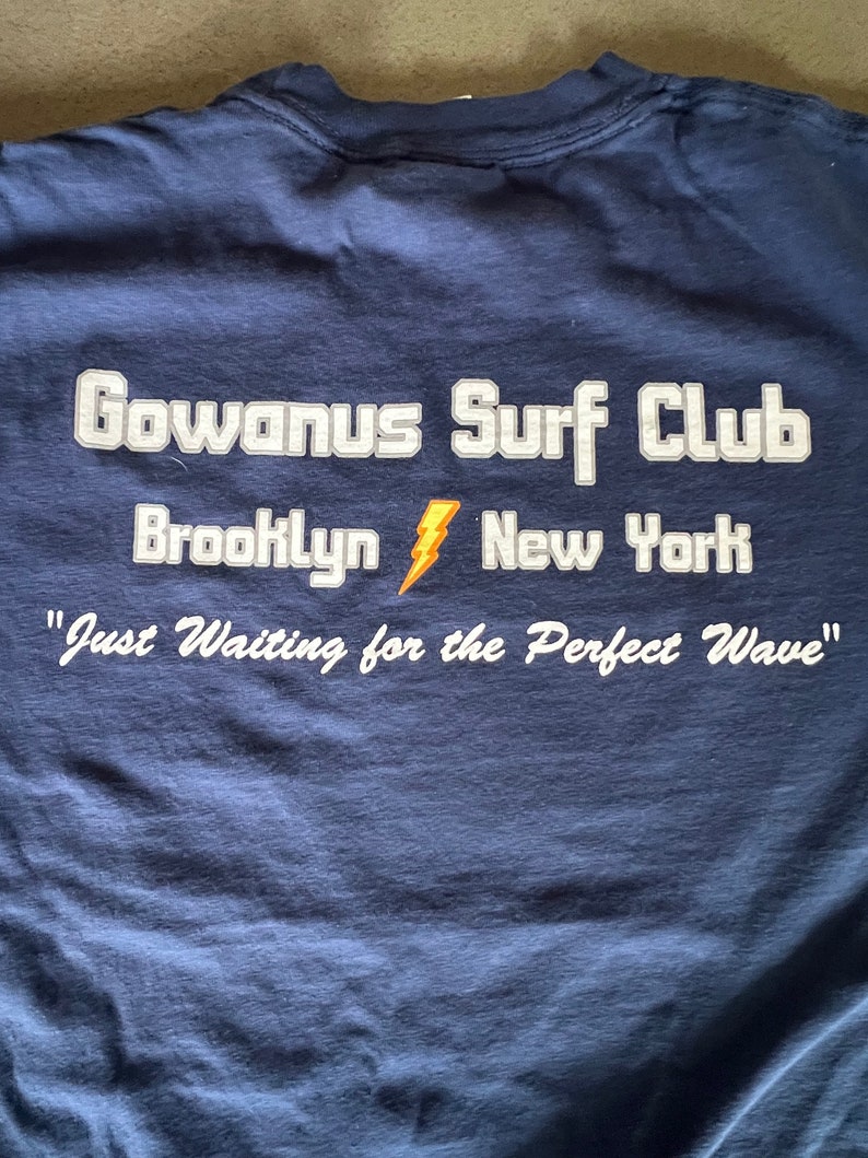 May include: A navy blue t-shirt with white text that reads "Gowanus Surf Club Brooklyn New York" and "Just Waiting for the Perfect Wave".