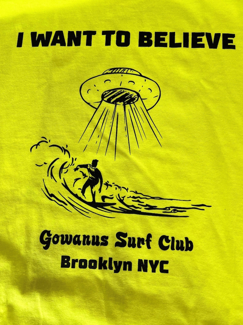 May include: Bright yellow t-shirt with the text "I WANT TO BELIEVE" above a graphic of a UFO beaming down on a surfer. Below the graphic is "Gowanus Surf Club Brooklyn NYC" in black text.