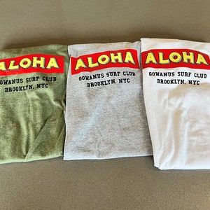 以下が含まれることがあります： 白い背景に赤と黒の文字で「ALOHA GOWANUS SURF CLUB BROOKLYN, NYC」と書かれた、折り畳まれたTシャツ3枚。Tシャツは緑、グレー、白です。