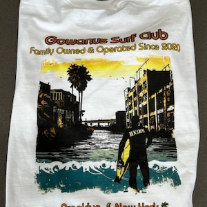 以下が含まれることがあります： 背景に建物が建ち並ぶ運河に立つサーファーのグラフィックプリントが施された白いTシャツ。Tシャツには「Gowanus Surf Club Family Owned & Operated Since 2020 Brooklyn New York」の文字がプリントされています。