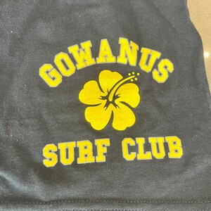 May include: Black t-shirt with a yellow hibiscus flower and the text "GOWANUS SURF CLUB" in yellow letters.