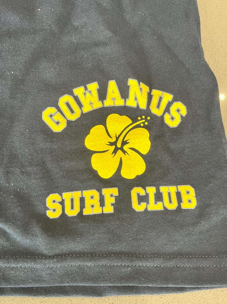 May include: Black t-shirt with a yellow hibiscus flower and the text "GOWANUS SURF CLUB" in yellow letters.