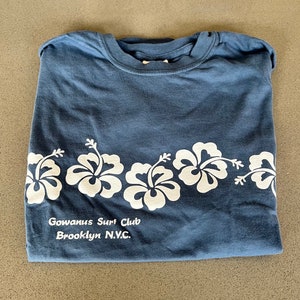 May include: A blue t-shirt with a white hibiscus flower design and the text "Gowanus Surf Club Brooklyn N.Y.C."
